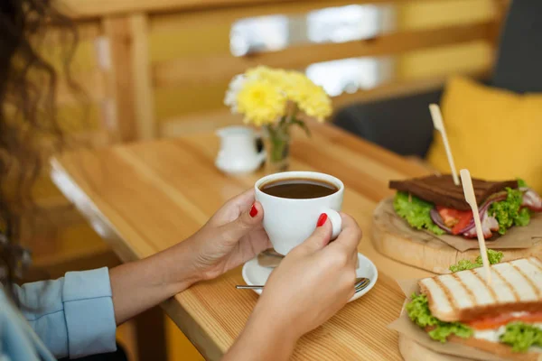 Vrouw houd van een kopje koffie aan backgroud houten tafel, waar een sandwich ligt — Stockfoto