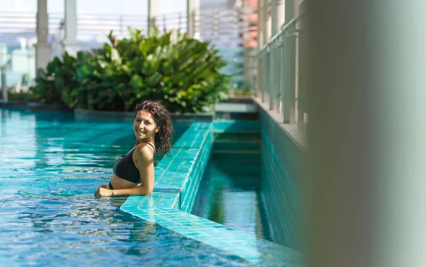 Πορτρέτο του ένα σέξι χαμογελαστό καυκάσιος γυναίκα σε ένα μαγιό χαλαρώνοντας στην πισίνα στον τελευταίο όροφο με πράσινο θάμνους και θέα στην πόλη. Ασία το Σαββατοκύριακο — Φωτογραφία Αρχείου