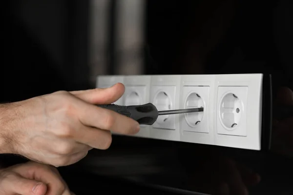 Close-up of an electrician\'s hand with a screwdriver disassembling a white electrical outlet on a black glass wall