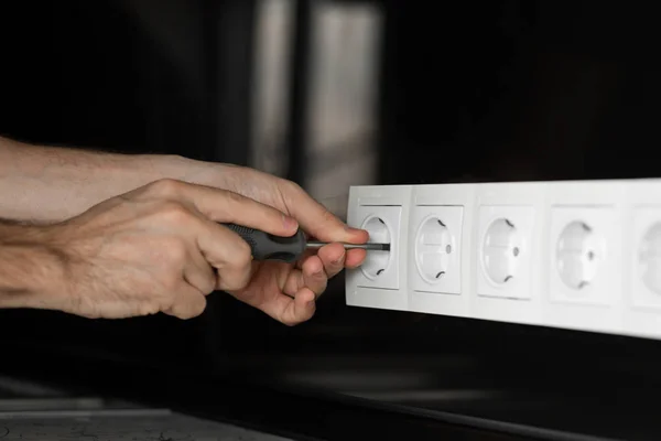 Close-up of an electrician\'s hand with a screwdriver disassembling a white electrical outlet on a black glass wall