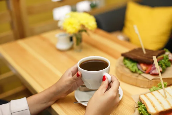 Vrouw houd van een kopje koffie aan backgroud houten tafel, waar een sandwich ligt — Stockfoto