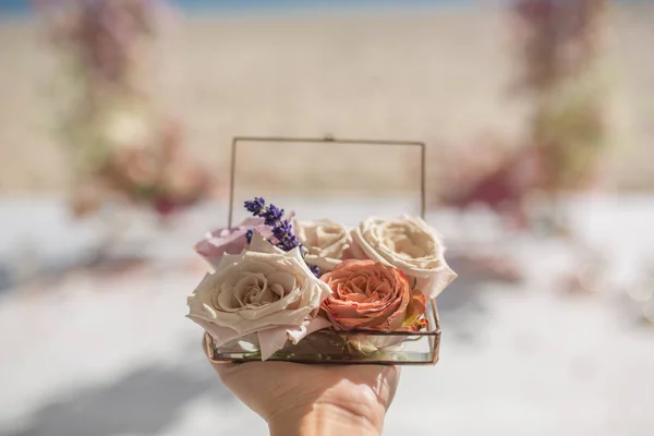 closeup woman hands hold glass box for wedding rings decorated with fresh rose flowers and banch of lavender at blurred coast background with wedding arch. Event decoration with fresh flowers