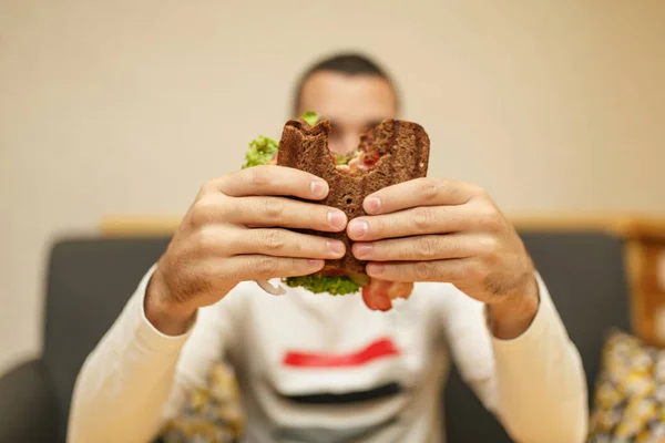Nahaufnahme witzig verschwommener Ausschnitt eines jungen Mannes, der ein angebissenes Sandwich mit beiden Händen hält. Sandwich im Fokus. heller Hintergrund. — Stockfoto