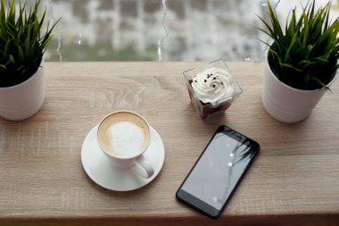 Beyaz daire, siyah cep telefonu, kırmızı kadife tatlı ve ahşap bar masa pencere yanında tencere yeşil çim üzerinde sıcak cappuccino beyaz fincan. Dışarıda soğuk karlı havalarda