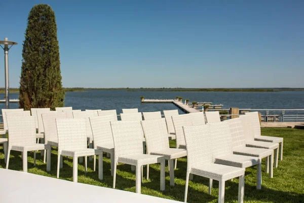 Schöne ausgehende Hochzeit eingerichtet. Romantische Hochzeitszeremonie, Hochzeit im Freien auf dem Rasen Wasserblick. Hochzeitsdekor. Weiße Holzstühle auf einem grünen Rasen. Weiße Sessel für Gäste — Stockfoto