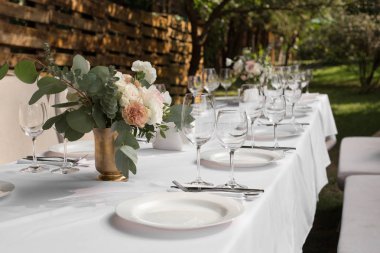 Pirinç bir vazoda taze çiçeklerle süslenmiş düğün masası. Düğün çiçekçisi olarak. Dışarıdaki yeşil manzaralı misafirler için bir ziyafet masası. Güllü buket, Eustoma ve okaliptüs yapraklı.