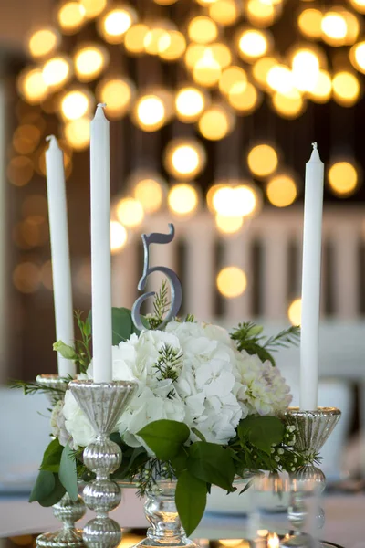 Bruiloft tafel setting is versierd met verse bloemen en witte kaarsen. Bruiloft bloemisten. Boeket met rozen, hortensia en eustoma. Op de achtergrond wazig branden bloemenslingers met gloeilampen. — Stockfoto