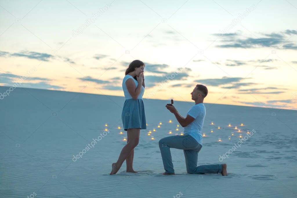 The guy makes the girl a marriage proposal by bending his knee while standing on the sand in the desert. Evening, candles burn in the sand