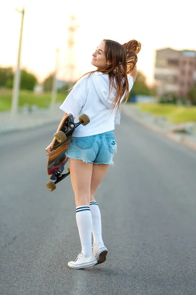 Street portrait of a girl in casual clothes and longboard in her hand stands on the playground, poses. Girl model with ride board posing at camera. Longboarding.
