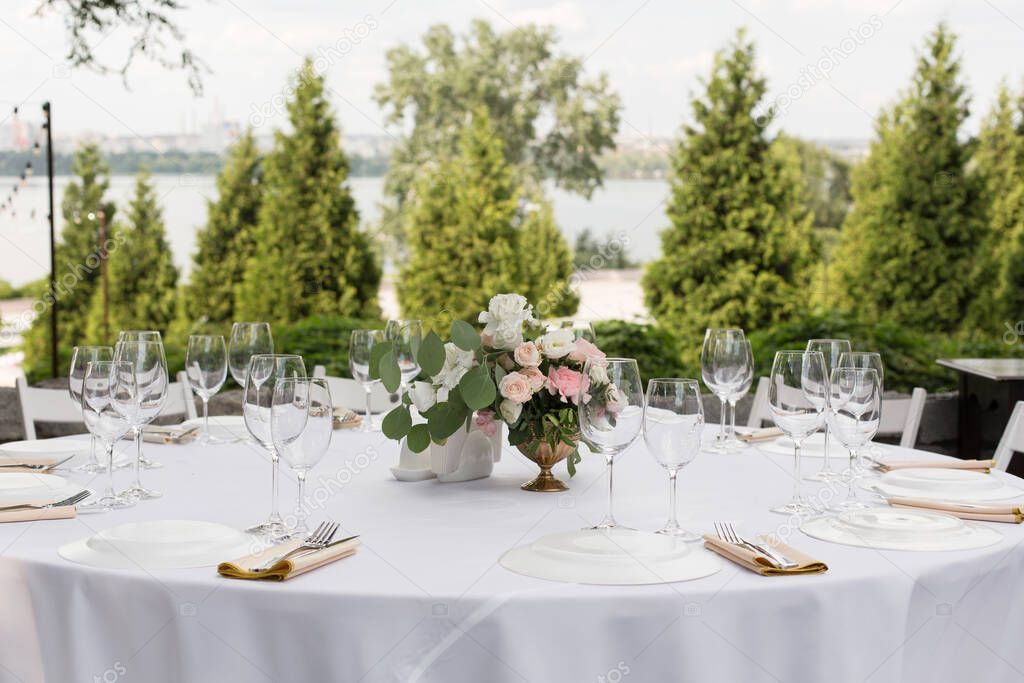 Wedding table setting decorated with fresh flowers in a brass vase. Wedding floristry. Banquet table for guests outdoors with a view of green nature. Bouquet with roses, eustoma and eucalyptus leaves.