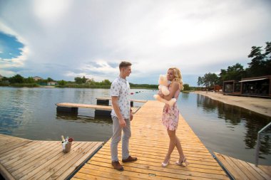 Romantik randevu sürprizi. Bir genç adam ve bir kız göle bakan bir iskelede. Adam kıza bir oyuncak ayı veriyor.