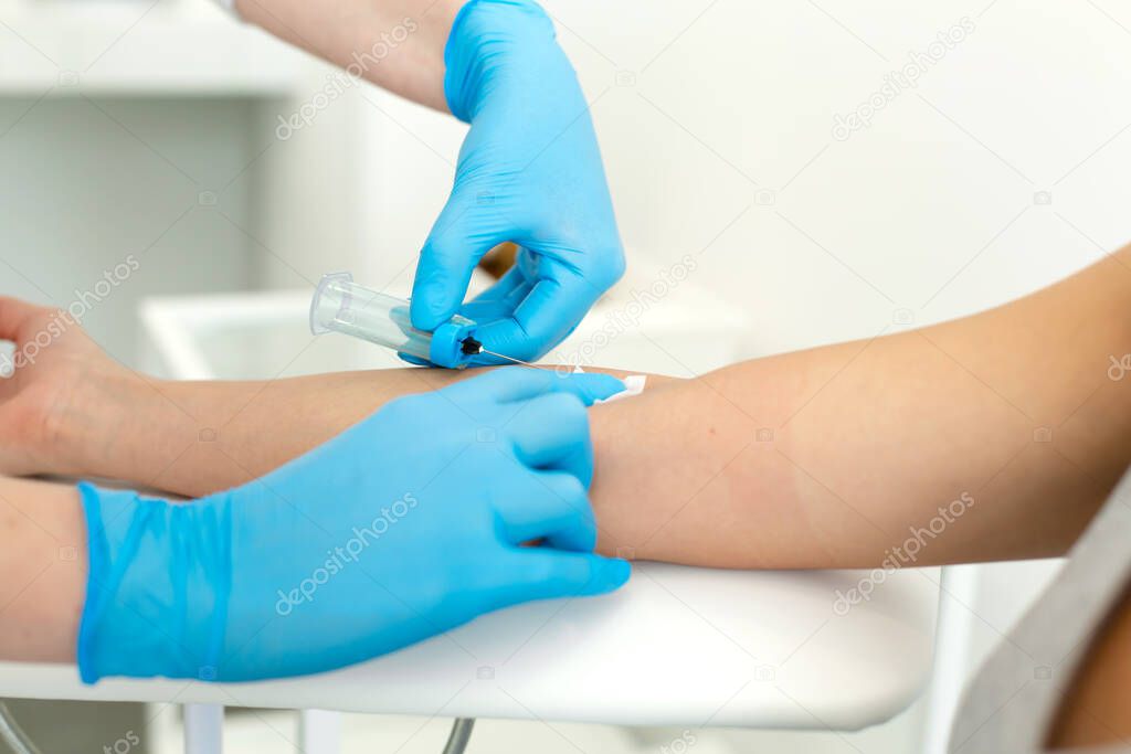 a gloved nurse inserts a needle into a vein on the patients arm and draws blood into a vacuum container. Blood sampling procedure.