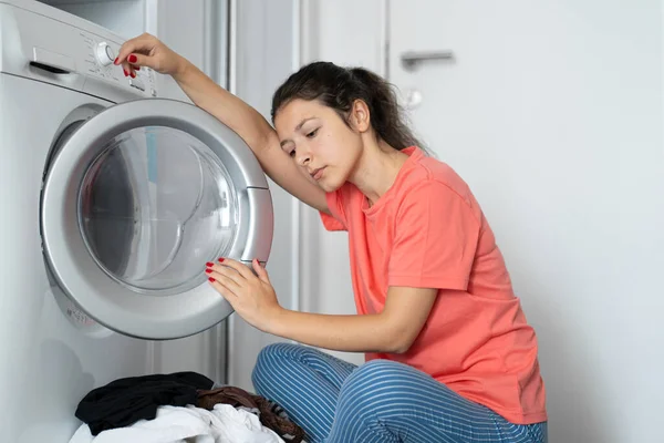 A girl loads dirty laundry into a washing machine while sitting on the floor in an apartment. Laundry day, housework.