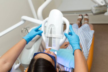 Diş beyazlatma cihazı ile diş beyazlatma, hastaya bakan diş asistanı, gözlük ile korunan gözler. Işık, lazer, florür ile beyazlatma. Yapay diş beyazlatma.