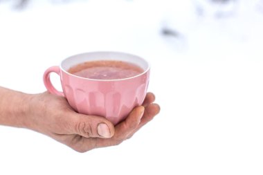 Bir kış manzarasının arka planında içkisi olan pembe bir kupayı tutan eller..