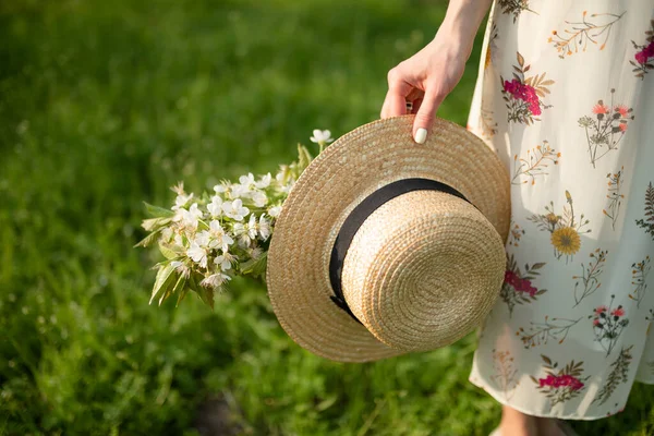 春の緑の公園に咲く自然を楽しむ夏の日差しの中を歩く少女は ウィッカーの帽子と花を手に持っています — ストック写真