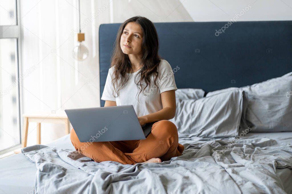 Beautiful young brunette girl with a laptop sitting on the bed. Stylish modern interior. A cozy workplace. Shopping on the Internet.
