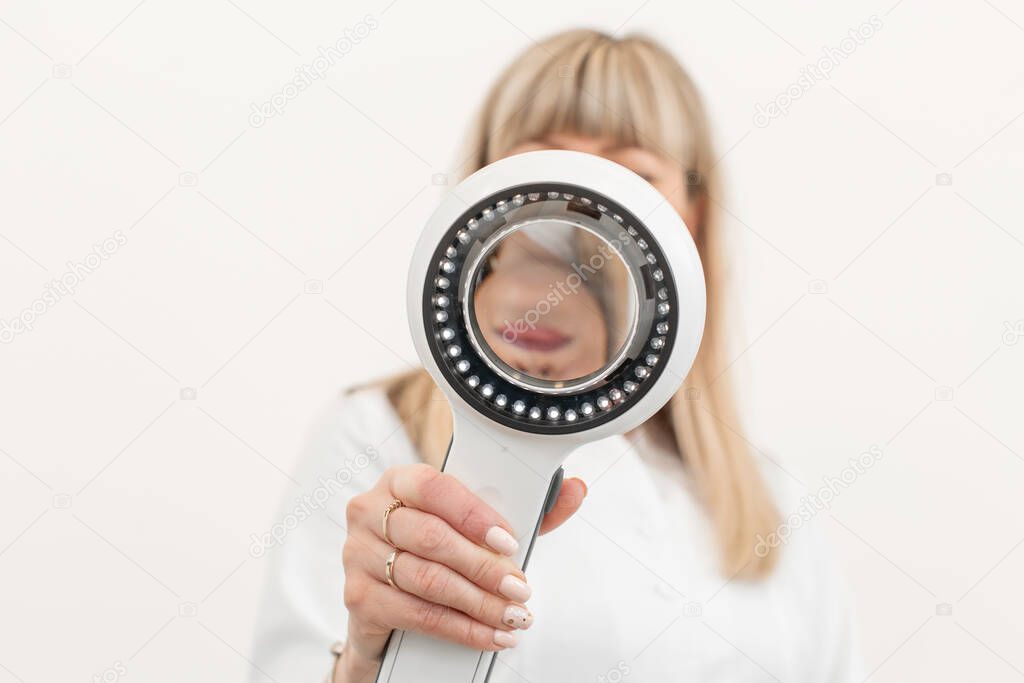 Portrait of a doctor trichologist dermatologist looking at a dermatoscope. Focus on the hand with a dermatoscope, the doctor is blurred. In a bright cosmetology room.