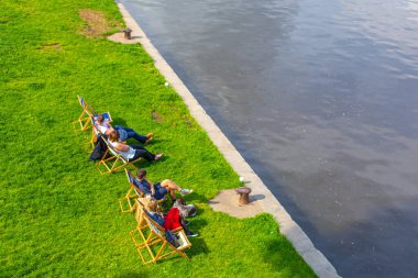 Üst manzara, insanlar nehir kenarındaki çimenlikteki tahta sandalyelerde dinleniyor. Prag / Çek Cumhuriyeti - 05.21.2019