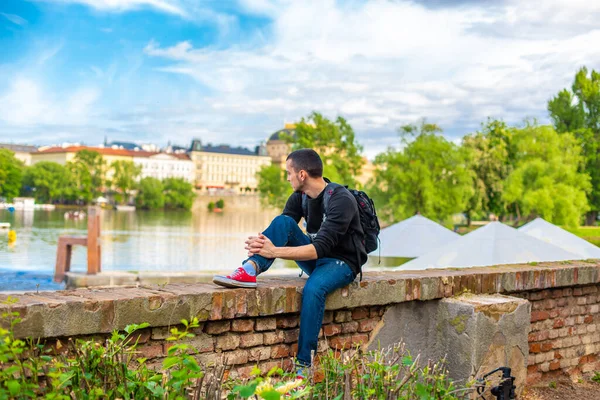 バックパックを持った観光客がヴルタヴァ川を見下ろすプラハの堤防に座っています — ストック写真