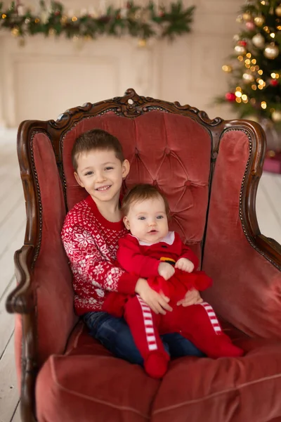 Litttle brother hugs his little sister in red retro chair near Christmas Tree. Enjoying a love hug, people\'s holidays. Togetherness concept.