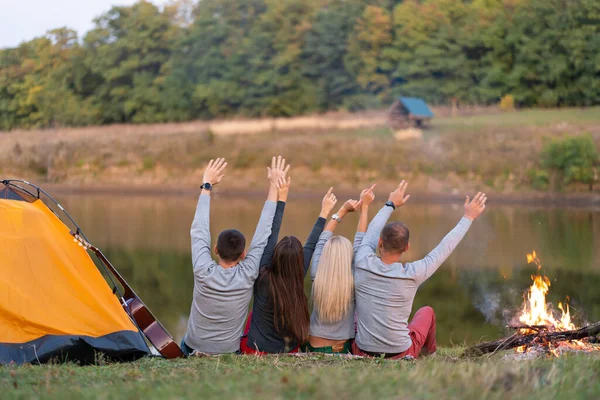 Shoot from back. A group of happy friends camping at riverside, dancing hold hands up and enjoy view. Holidays fun.