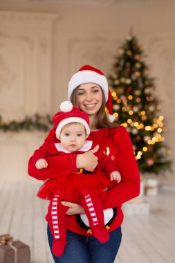Mutlu anne, Noel Baba kostümlü ve şapkalı küçük kızı Noel ağacının yanında kucakla..