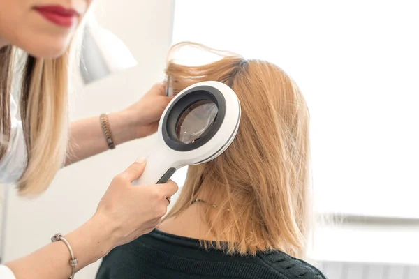 Bir tricholog, hastaların başlarındaki kılların durumunu dermatoskopla inceliyor. Parlak bir kozmetoloji odasında.