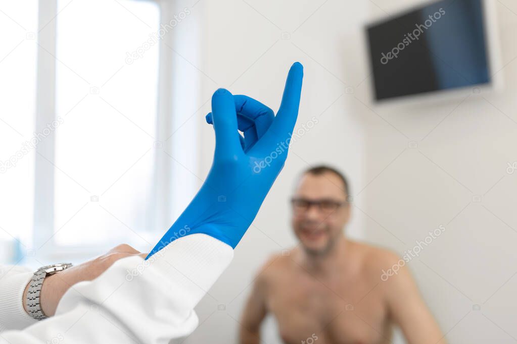 urologist doctor puts on medical gloves before examining a patient who is waiting on a couch.