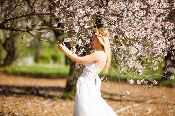 年轻美丽的浪漫金发碧眼的女人在白色礼服采取自拍在盛开的花园 — 图库照片