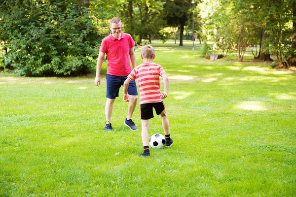 快乐的年轻父亲和他的小儿子在阳光明媚的绿色公园踢足球 — 图库照片