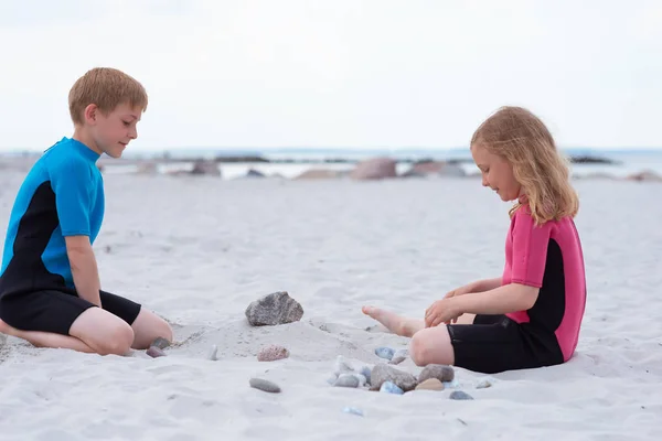 Dos niños en trajes de baño de neopreno jugando en la playa con san —  Fotos de Stock