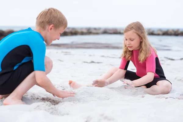 Zwei Kinder in Neopren-Badeanzügen spielen am Strand mit san — Stockfoto