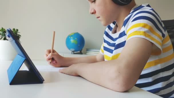 十代の男の子は タブレットやヘッドフォンを使用してビデオレッスンを持っており ホームスクールで鉛筆で通知を書きます — ストック動画