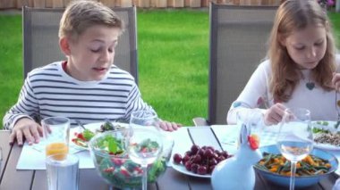 İki mutlu genç çocuk, aileleriyle sohbet edip yemek yiyorlar. Sıcak arka bahçenin terasında sağlıklı bir akşam yemeği yiyorlar.