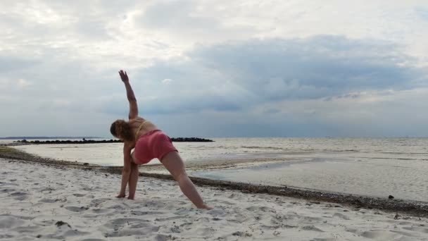 4K率直なビデオの美しいです若いです女性作るヨガAsanasオンザビーチで日没時バルト海 — ストック動画