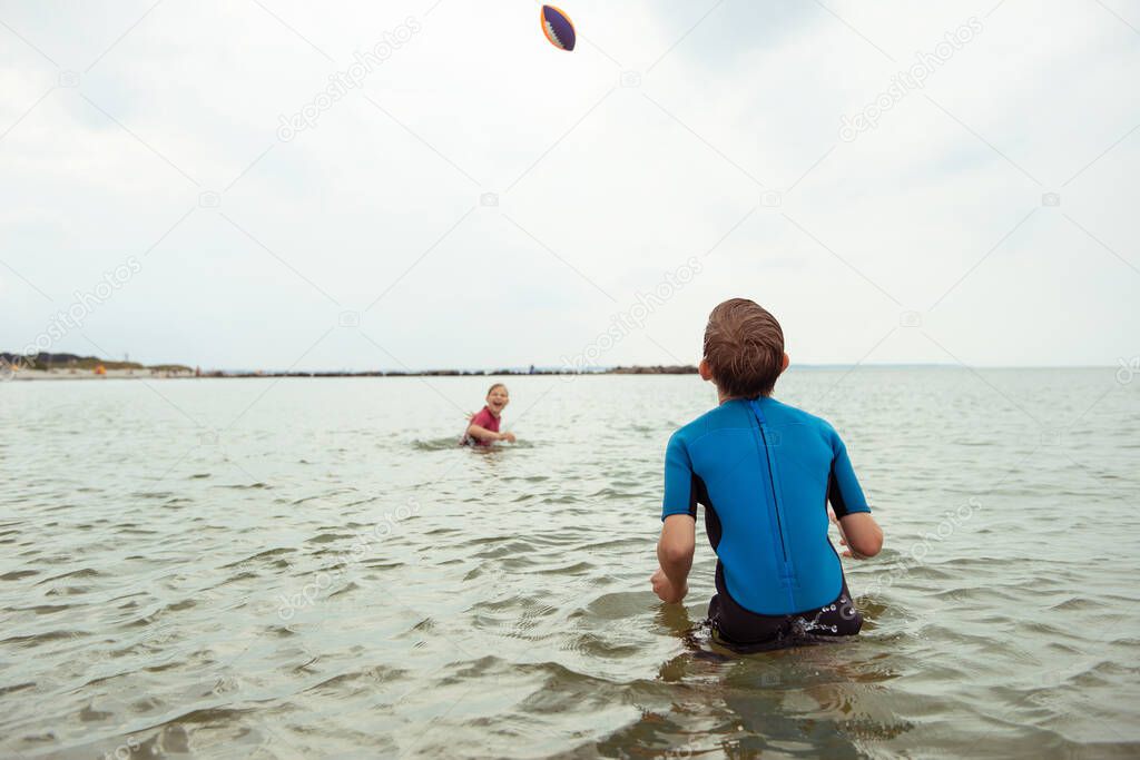 Two happy siblings children playing and jumping with ball in water in neoprene suits in Baltic sea