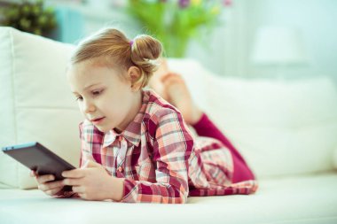 Dijital ev okulunda elinde tabletle kanepede yatan sevimli küçük kız çocuğu portresi.