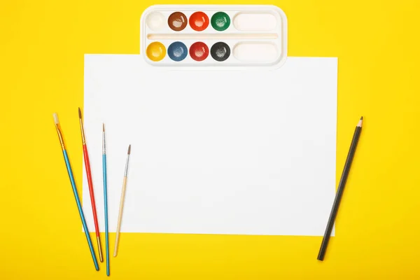 Blank paper with pencil, brushes and paints on yellow background. Artist workplace concept with copy space for your ideas. — Stock Photo, Image