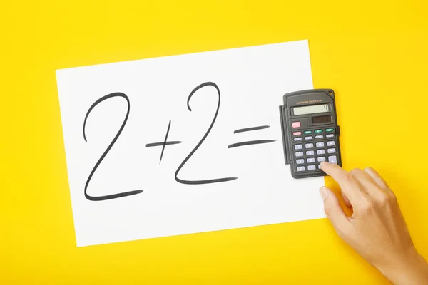 Papel em branco com tarefa 2 mais 2 e com uma solução em calculadora com mão sobre fundo amarelo — Fotografia de Stock