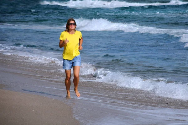 漂亮的白种人年轻的微笑的女人 头发蓬松 穿着黄色的T恤和蓝色的磨损短裤 沿着海滩奔跑 — 图库照片