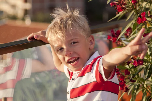 Blond boy looks at the camera and throws his hand up with a vict — Stock Photo, Image