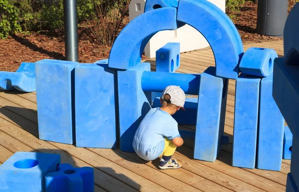 Little boy is sitting on childrens playground in park with blue giant geometric figures for the development of imagination and spatial thinking — Stock Photo, Image