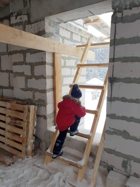 Χαρούμενο αγόρι με κόκκινο σακάκι σκαρφαλώνει ξύλινη σκαλωσιά στο παράθυρο του σπιτιού υπό κατασκευή το χειμώνα — Φωτογραφία Αρχείου