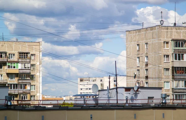 Satellite dishes and power line wires on metal roof of residential building against background of other multistory residential panel buildings and blue sky. A lot of wires on roof of house