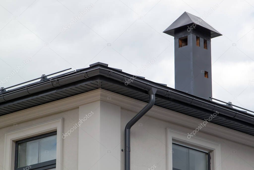 close-up view of house with gray fold roof and plums and filing of roof overhangs with soffits and brick stone pipe covered with metal sheets and black smoke box