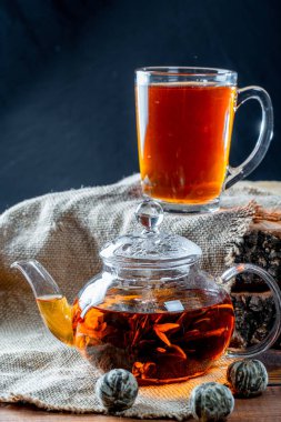 Çiçeklenme büyük çiçekli bir cam demlik çay. Egzotik yeşil çay-topları çiçek çiçek ve bir fincan bardak çay çaydanlık. Çuval bezi ve eski ahşap güdük arka plan üzerinde çay töreni