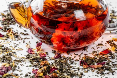 Çiçeklenme büyük çiçekli bir cam demlik çay. Dağınık bir beyaz zemin üzerine egzotik yeşil çay demlik çay yaprakları ve meyve parçaları kurutulmuş