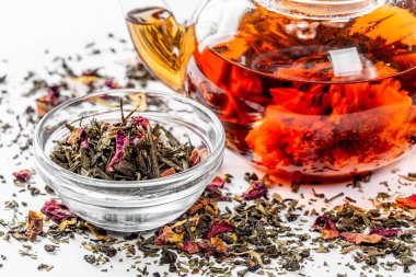 Çiçeklenme büyük çiçekli bir cam demlik çay. Dağınık bir beyaz zemin üzerine egzotik yeşil çay demlik çay yaprakları ve meyve parçaları kurutulmuş