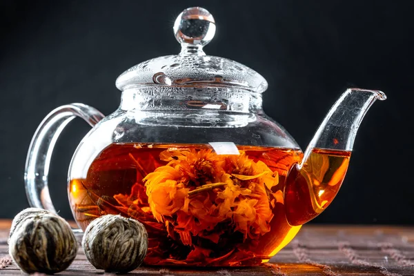 Tea in a glass teapot with a blooming large flower. Teapot with exotic green tea-balls blooms flower. Tea ceremony on a dark background